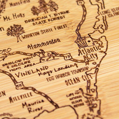 A detailed view of the "A Slice of Life New Jersey Cutting Board" by Totally Bamboo reveals an artfully engraved map of southern New Jersey. This intricately designed cutting board spotlights locations such as Atlantic City, Hammonton, Vineland, and Ocean City and features various towns, highways, and natural areas in an elegant state outline design. The board measures 11 inches by 8-3/4 inches.