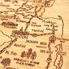 A detailed photo of the "A Slice of Life New Jersey Cutting Board" by Totally Bamboo, featuring intricate engravings of New Jersey's cities and towns like Trenton, Freehold, and Asbury Park. This wooden board captures roads and landmarks in a vintage-style design with a rustic aesthetic.