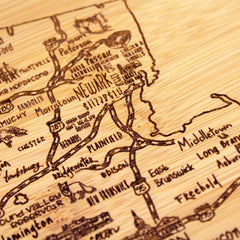 A close-up view of the "A Slice of Life New Jersey Cutting Board" by Totally Bamboo, showcasing a wooden map engraved with the names and routes of New Jersey towns such as Newark, Middletown, and Freehold. The intricately detailed roads and highways provide a rustic and artistic depiction of the area.