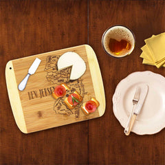 On a wooden table sits the "A Slice of Life New Jersey Cutting Board" by Totally Bamboo, measuring 11" x 8-3/4", engraved with the outline of New Jersey and holding a cheese knife, cheese, and crackers topped with tomato and basil. Nearby are a glass of beer, yellow napkins, and a white plate with silverware.