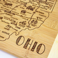 The Totally Bamboo "A Slice of Life Ohio Cutting Board," measuring 11" x 8-3/4", serves as both a practical charcuterie board and a detailed map of Ohio, complete with city names and illustrations of landmarks. The word "OHIO" is prominently etched at the bottom, making it a functional piece and unique artwork.