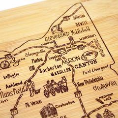 A detailed view of the laser-engraved design on the A Slice of Life Ohio Cutting Board by Totally Bamboo showcases cities such as Akron, Canton, Cleveland, Medina, and Massillon. This charcuterie board features intricately etched roads, historical figures, and landmarks on a light wooden surface.