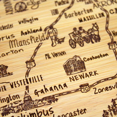 The A Slice of Life Ohio Cutting Board by Totally Bamboo features laser-engraved artwork depicting a wooden map with hand-drawn illustrations and labels of cities such as Columbus, Mansfield, Newark, and Zanesville in Ohio. It also includes decorative sketches of historical scenes and landmarks seamlessly incorporated into the design.