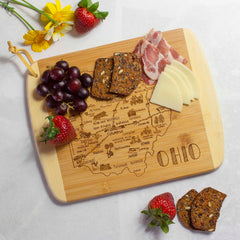 A charcuterie board from Totally Bamboo's *A Slice of Life Ohio* collection, featuring laser-engraved artwork of Ohio's map with city names, elegantly displays assorted snacks such as crackers, cheese slices, salami, red grapes, and strawberries. Yellow and white flowers are beautifully arranged beside the 11" x 8-3/4" bamboo cutting board on a white surface.