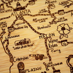 The 11" x 8-3/4" "A Slice of Life Georgia Cutting Board" by Totally Bamboo is a meticulously detailed wooden map of Georgia, designed like a charcuterie board. It highlights cities such as Atlanta, Macon, and Columbus and showcases notable landmarks like the "Little White House" and various lakes. Hand-drawn illustrations and routes contribute to the rustic, artistic charm of this distinctive state outline.