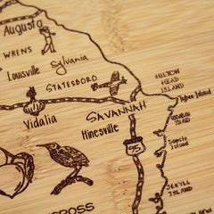 Close-up of the "A Slice of Life Georgia Cutting Board" by Totally Bamboo, laser-engraved to display southeastern Georgia, highlighting cities such as Savannah and Hinesville. The design features illustrations like a bird, an airplane, and Interstate 95, all framed within the unique outline of the state.