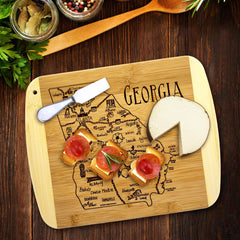 A Totally Bamboo "A Slice of Life Georgia" cutting board, measuring 11" x 8-3/4", showcases the state outline of Georgia with a round cheese wheel and three bread slices adorned with salami and rosemary. A butter knife rests on the charcuterie board, surrounded by various herbs and condiments on a wooden table.