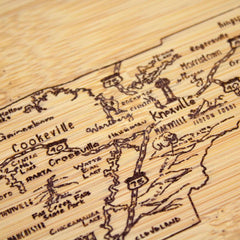 A close-up of a laser-engraved artwork showcases the Totally Bamboo "A Slice of Life Tennessee Cutting Board," featuring a wooden map that highlights locations such as Knoxville, Cookeville, and Crossville. Roads and interstates like I-40 and I-75 are intricately detailed on this stunning 11" x 8-3/4" piece.