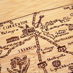 The "A Slice of Life Tennessee Cutting Board" by Totally Bamboo showcases a laser-engraved design on bamboo, highlighting routes and landmarks around Nashville. It features road numbers, natural elements, and artistic renditions of buildings and flowers, making it an ideal decorative charcuterie board. The dimensions are 11" x 8-3/4".