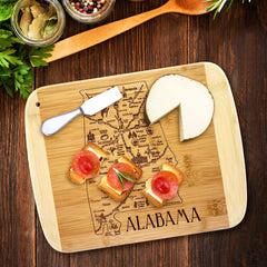 As part of Totally Bamboo's Slice of Life Collection, the A Slice of Life Alabama Cutting Board (11" x 8-3/4") features a detailed map design engraved into its bamboo surface. It's perfect for elegantly presenting cheese, a spreader, and small bread pieces with toppings, all beautifully surrounded by fresh herbs and a jar of peppercorns on the wooden table.