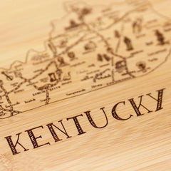 A close-up of the Totally Bamboo "A Slice of Life Kentucky Cutting Board," measuring 11" x 8-3/4", shows the state outline artwork of Kentucky, reminiscent of a bamboo cutting board. The word "Kentucky" is prominently engraved in bold letters below, highlighting its notable landmarks as part of the collection.