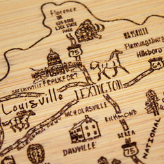 A close-up of the Slice of Life Kentucky Cutting Board by Totally Bamboo, measuring 11" x 8-3/4", showcases a hand-drawn map featuring towns such as Louisville, Lexington, and Frankfort in Kentucky. This state outline artwork includes depictions of animals, a building, and highways in a rustic, artistic style.