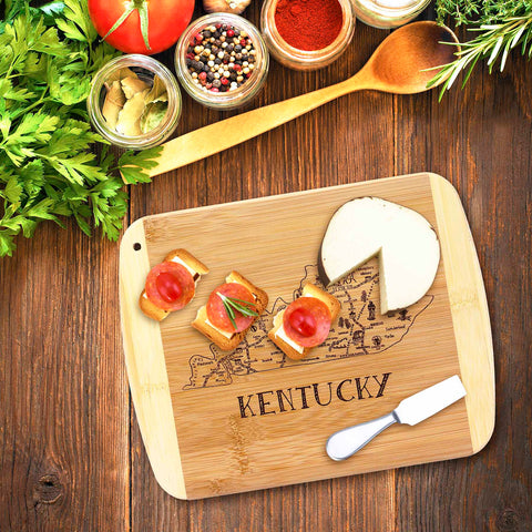 The Totally Bamboo A Slice of Life Kentucky Cutting Board, measuring 11" x 8-3/4", features the engraved state outline artwork of Kentucky and is showcased holding a round cheese wheel and snacks. Nearby lies a small knife, surrounded by fresh herbs, spices, and a tomato on a wooden table.