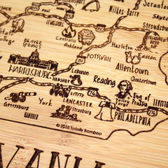 The "A Slice of Life Pennsylvania Cutting Board" by Totally Bamboo is a charming wooden piece that depicts the state in an illustrative style, highlighting cities like Philadelphia, Lancaster, and Reading with decorative icons and text. The laser-engraved artwork adds detail to key places and roads on this attractive 11" x 8-3/4" board.