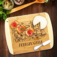 An "A Slice of Life Pennsylvania Cutting Board" by Totally Bamboo, measuring 11" x 8-3/4", features a bamboo surface laser-engraved with a map of Pennsylvania and the word "Pennsylvania." It elegantly holds three snacks with round toppings and a wedge of cheese. Nearby are herbs, spices, a spoon, and a fork, creating the perfect charcuterie-serving experience.