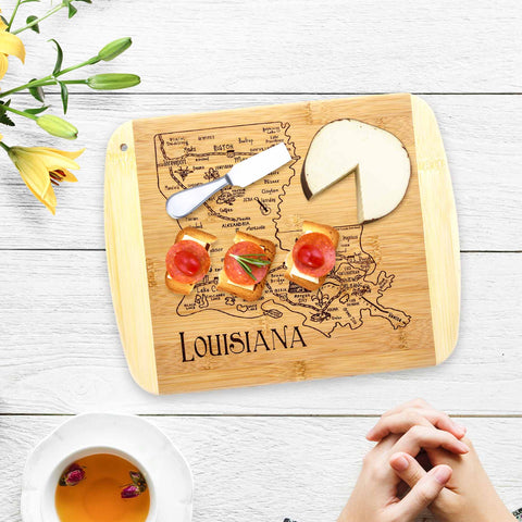 A cutting board from Totally Bamboo's A Slice of Life Collection features the engraved outline of Louisiana and showcases round cheese and three crackers adorned with tomatoes and herbs. A nearby bouquet and steaming cup of tea sit on a white surface, while hands gently clasp beneath.