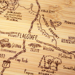 The "A Slice of Life Arizona Cutting Board" by Totally Bamboo is a carved wooden map that highlights Flagstaff, Arizona, and surrounding regions. This state-themed cutting board features detailed illustrations of the Grand Canyon, Painted Desert, and Meteor Crater, with roads and small icons showcasing local landmarks and attractions. It measures 11" x 8-3/4".