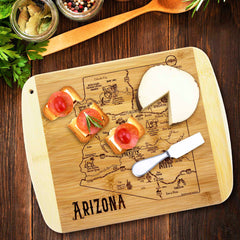 The "A Slice of Life Arizona Cutting Board" by Totally Bamboo elegantly showcases small appetizers and cheese, engraved with a map of Arizona. A cheese knife is conveniently placed beside it, while nearby jars of spices and a wooden spoon enhance the charm of this state-themed culinary experience.