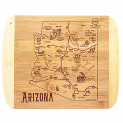 The "A Slice of Life Arizona" cutting board by Totally Bamboo, measuring 11" x 8-3/4", is made from wood and features a detailed map of Arizona engraved on it. This unique piece highlights cities, landmarks, and illustrations such as mountains, animals, and cacti, with the word "Arizona" elegantly placed in the bottom left corner.