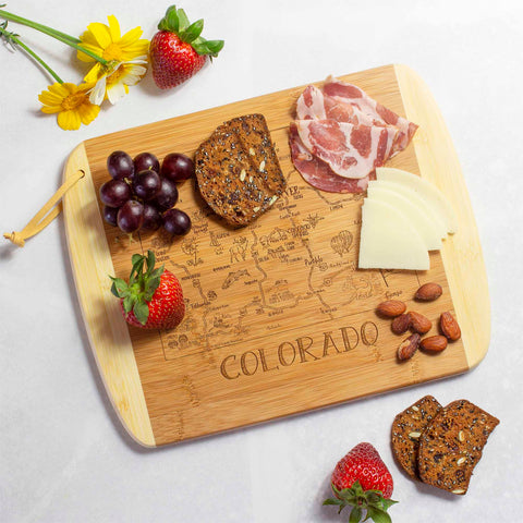 As a piece from the Slice of Life Collection by Totally Bamboo, the A Slice of Life Colorado Cutting Board (11" x 8-3/4") is beautifully engraved with "Colorado." Showcased on a white surface, it elegantly holds round meat slices, cheese wedges, seeded crackers, almonds, strawberries, grapes, daisies, and other artfully arranged items.