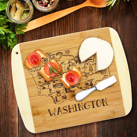 The "A Slice of Life Washington Cutting Board" by Totally Bamboo, measuring 11" x 8-3/4", showcases an engraved map of Washington state. It elegantly holds a block of cheese, a cheese knife, and three small toast slices garnished with cheese, tomato, and rosemary. Nearby lay a wooden spoon alongside herbs and spices.