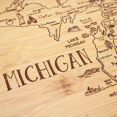 The "A Slice of Life Michigan Cutting Board" by Totally Bamboo, measuring 11" x 8-3/4", is meticulously crafted in the shape of Michigan's state outline, showcasing intricate details of towns, regions, and Lake Michigan. This charcuterie serving piece prominently features the word "MICHIGAN" elegantly engraved across its surface.