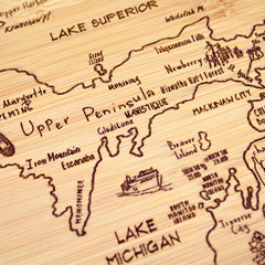 Close-up of the Totally Bamboo "A Slice of Life Michigan" Cutting Board (11" x 8-3/4"), crafted from wood and resembling a charcuterie board. It features detailed engravings of Michigan's Upper Peninsula with landmarks such as Lake Superior, and cities like Marquette, Munising, and Escanaba, along with a small boat illustration.