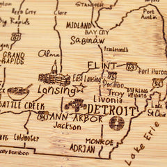 The "A Slice of Life Michigan Cutting Board" by Totally Bamboo, measuring 11" x 8-3/4", intricately displays a carved wooden map of Michigan, highlighting cities such as Grand Rapids, Saginaw, Flint, Lansing, Ann Arbor, and Detroit. This unique piece includes major highways and distinctive illustrations that represent each area.