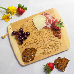 A charcuterie board, called "A Slice of Life Michigan Cutting Board" by Totally Bamboo, measuring 11" x 8-3/4", features an outline of Michigan. It is crafted from bamboo and beautifully presents a selection of cheese, cured meats, strawberries, grapes, and almonds. Nearby are crackers and a strawberry complemented by vibrant yellow flowers on a pristine white surface.