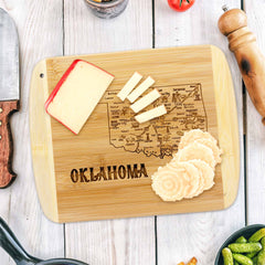 The "A Slice of Life Oklahoma Cutting Board," measuring 11" x 8-3/4" by Totally Bamboo, is engraved with an Oklahoma state outline artwork and elegantly arranged with sliced cheese and crackers. A knife, pepper grinder, and various ingredients are scattered around it on a light wooden table.