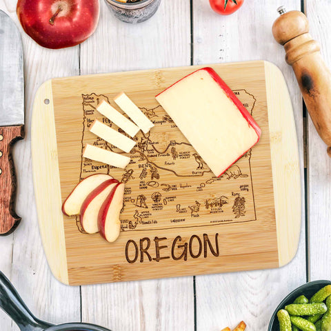 The Totally Bamboo A Slice of Life Oregon Cutting Board, measuring 11" x 8-3/4", features a map of Oregon and beautifully arranged slices of cheese and apple. As part of the Slice of Life Collection, this state outline artwork is paired with kitchen staples such as a pepper grinder, knife, and fresh vegetables.