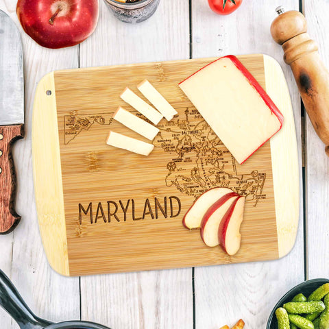 The "A Slice of Life Maryland Cutting Board" by Totally Bamboo, measuring 11" x 8-3/4", showcases a Maryland state outline engraving. It features cheese slices and apple wedges, complementing an arrangement with an apple, pepper mill, knife, and assorted vegetables on a rustic wooden table.