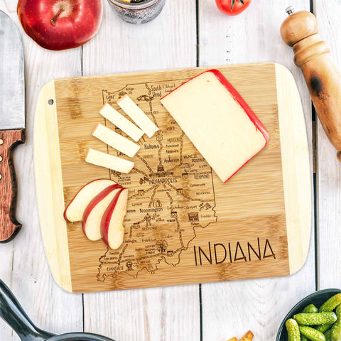 From Totally Bamboo's A Slice of Life Collection, the Indiana Cutting Board, measuring 11" x 8-3/4", is featured on the kitchen table. It is elegantly decorated with slices of cheese and apple, while whole fruits, a knife, and a pepper grinder surround it.