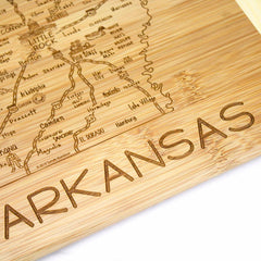 The Totally Bamboo "A Slice of Life Arkansas Cutting Board" measures 11" x 8-3/4" and showcases a detailed engraving of a map of Arkansas. The board features city names such as Little Rock and El Dorado, with "ARKANSAS" prominently carved into the bottom right corner, making it an ideal piece of artwork for any kitchen.