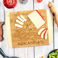 This 11" x 8-3/4" A Slice of Life Arkansas Cutting Board by Totally Bamboo showcases an engraved map of Arkansas. It's elegantly topped with sliced cheese and apple, accompanied by a knife, whole apple, pepper grinder, and various vegetables, all artfully displayed on a white wooden surface.