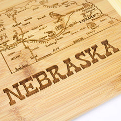 From the Slice of Life Collection by Totally Bamboo, the A Slice of Life Nebraska Cutting Board (11" x 8-3/4") is crafted from light natural wood and designed as a charcuterie serving board. It showcases an engraved map of Nebraska, featuring key cities and landmarks, with "Nebraska" prominently etched at the bottom.