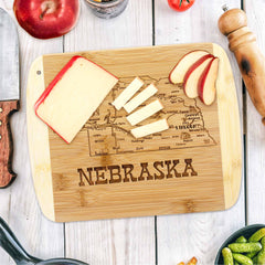 From the Slice of Life Collection by Totally Bamboo, this delightful "A Slice of Life Nebraska Cutting Board" measures 11" x 8-3/4" and showcases an intricately engraved map of Nebraska. It elegantly holds slices of cheese and apple, complemented by a knife, pepper mill, an apple, and fresh vegetables on a white wooden table.