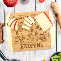 This cutting board from Totally Bamboo's "A Slice of Life" collection is laser-engraved with a map of Wyoming adorned with local icons. It measures 11" x 8-3/4" and elegantly presents slices of cheese and apple alongside kitchen essentials such as a knife, pepper grinder, and tomato.