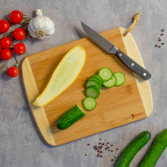 A sliced cucumber sits on the Totally Bamboo A Slice of Life Nevada Cutting Board, 11" x 8-3/4", along with a knife. Whole cucumbers, cherry tomatoes, garlic cloves, and scattered peppercorns are arranged around them on a gray surface. Part of the Slice of Life Collection, this scene beautifully encapsulates the essence of fresh ingredients poised for culinary artistry.