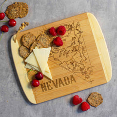 The Totally Bamboo "A Slice of Life Nevada Cutting Board, 11" x 8-3/4" is beautifully engraved with the state outline of Nevada and showcases an arrangement of triangular slices of cheese, round whole-grain crackers, fresh raspberries, and cherries on a gray surface.