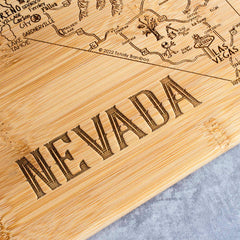 Close-up of the "A Slice of Life Nevada Cutting Board, 11" x 8-3/4"" from Totally Bamboo, featuring an intricately engraved map of Nevada. The prominently etched word "Nevada" stands out in large letters, with cities and landmarks like Reno and Las Vegas visible as the board rests on a gray surface.