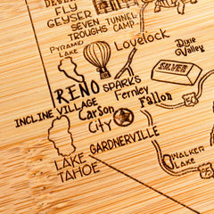 From the Slice of Life Collection by Totally Bamboo, the A Slice of Life Nevada Cutting Board (11" x 8-3/4") is crafted from wood to resemble a bamboo cutting board, featuring engraved depictions of Nevada's iconic locations such as Reno, Carson City, Lake Tahoe, and Walker Lake. It also showcases delightful illustrations of a hot air balloon and outdoor activities.