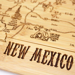 Close-up of the "A Slice of Life New Mexico Cutting Board" by Totally Bamboo, resembling a bamboo cutting board and featuring a laser-engraved design of New Mexico. The artwork highlights landmarks such as Carlsbad Caverns National Park, with "NEW MEXICO" prominently etched at the bottom.