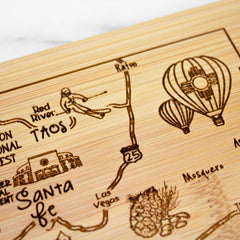 Introducing the A Slice of Life New Mexico Cutting Board by Totally Bamboo: an 11" x 8-3/4" bamboo cutting board exquisitely laser-engraved with a map featuring hot air balloons, a skier, and destinations such as Santa Fe, Taos, and Las Vegas. This design beautifully captures natural and cultural symbols through detailed sketches.