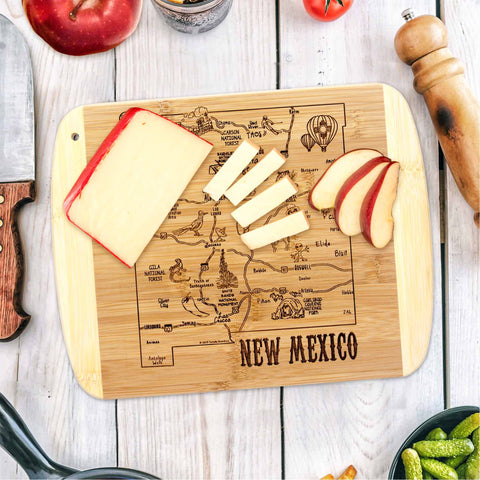 The A Slice of Life New Mexico Cutting Board by Totally Bamboo, measuring 11" x 8-3/4", features a laser-engraved map of New Mexico and adds elegance to serving cheese and apple slices. Accompanying this culinary centerpiece are a pepper shaker, half an apple, a tomato, and assorted vegetables.