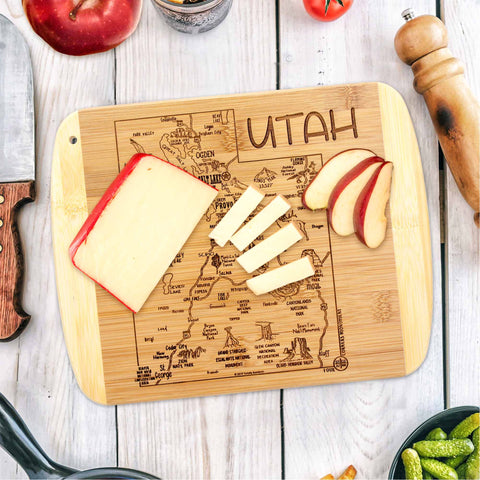 An A Slice of Life Utah Cutting Board by Totally Bamboo, measuring 11" x 8-3/4", features laser-engraved artwork of a Utah map and is adorned with cheese slices, a wedge of cheese, and apple slices. Accompanying the scene are a knife, a pepper grinder, a tomato, and a bowl of pickles on a light wooden table.