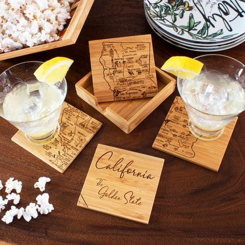 The table is adorned with the 4-Piece California Puzzle Coaster Set with Case by Totally Bamboo, featuring a laser-engraved map of California and the phrase "The Golden State." Two cocktails garnished with lemon slices sit atop these distinctive coasters. Nearby, a wooden dish overflows with popcorn, complemented by decorative plates to enhance the setting's charm.