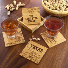 A pair of drinks sit on a 4-Piece Texas Puzzle Coaster Set with Case by Totally Bamboo, each featuring laser-engraved maps of Texas and the label "The Lone Star State." Accompanied by a wooden holder and a bowl of peanuts, they create a charming display on the wooden table, perfect for gifting to those who value distinctive craftsmanship.