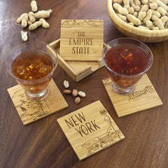 Two cocktails rest on a couple of coasters from the Totally Bamboo 4-Piece New York Puzzle Coaster Set with Case, each laser-engraved with "New York" and "The Empire State," adding a touch of charm to the wooden table. A bowl of peanuts is in the background, with scattered nuts enhancing the cozy setting.