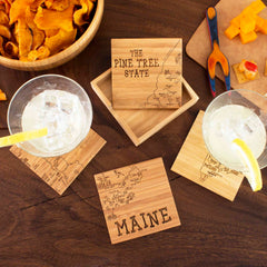 Two drinks with lemon slices sit on a wooden table, accompanied by a distinctive 4-Piece Maine Puzzle Coaster Set with Case from Totally Bamboo, engraved with "Maine" and "The Pine Tree State." Nearby are chips in a bowl and a plate featuring sliced cheese and crackers, alongside wooden utensils.